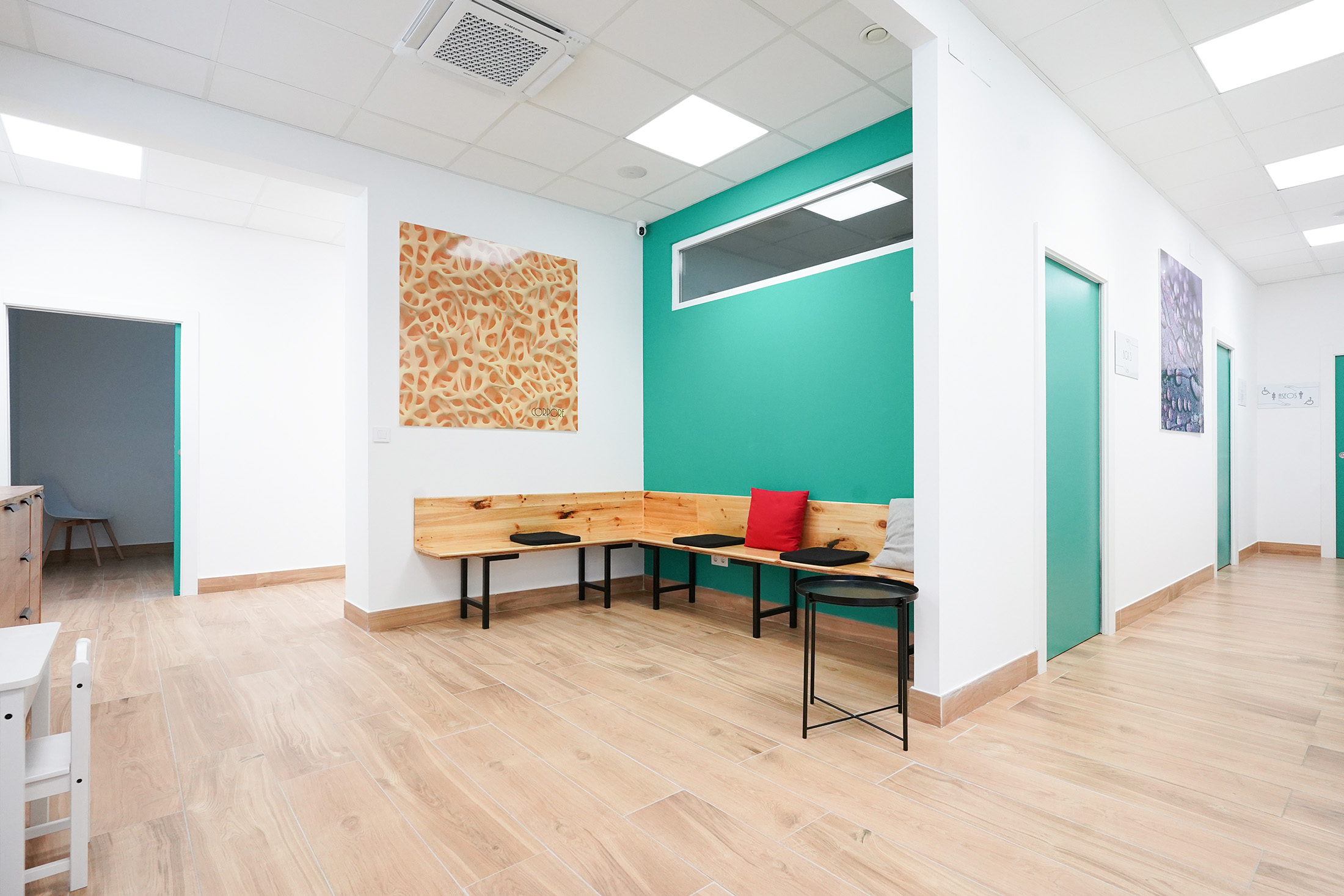 Sala de espera interior con suelo de madera, banco en forma de L y contraste de paredes blancas con pared de acento turquesa